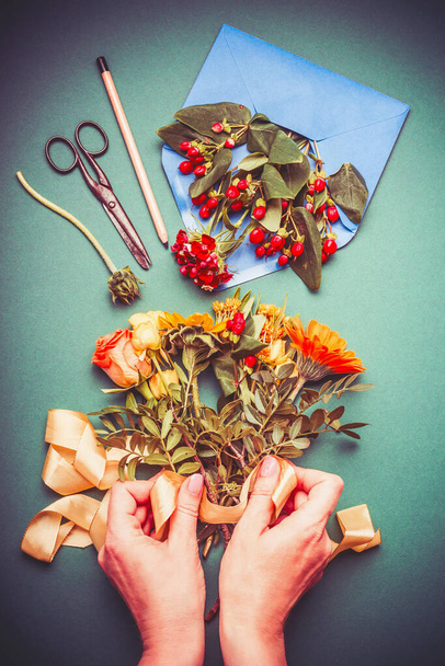 Female hand making autumn flowers arrangements on table workspace with scissors , envelope and ribbon, top view - Valokuva, kuva