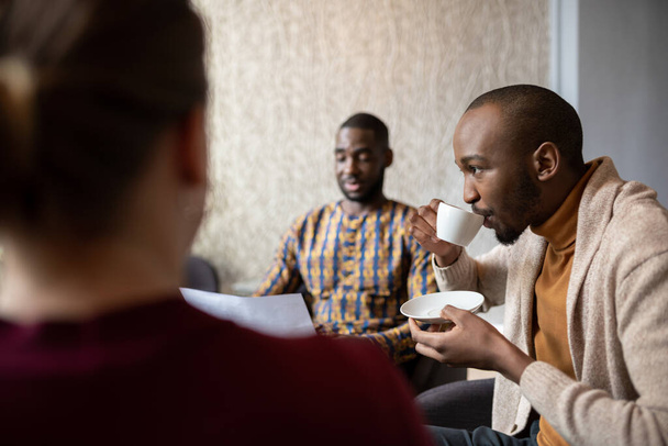 オフィスのラウンジエリアで同僚とのカジュアルなミーティング中にコーヒーを飲んでいる若いアフリカのビジネスマン - 写真・画像