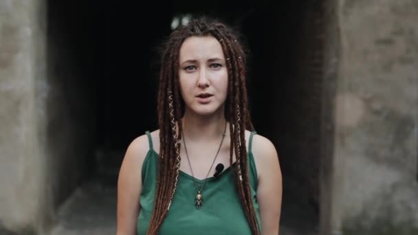 A pleasant young girl with dreadlocks and an interesting pendant around her neck leads a reportage standing in the background of a dark arch. Close-up - Footage, Video