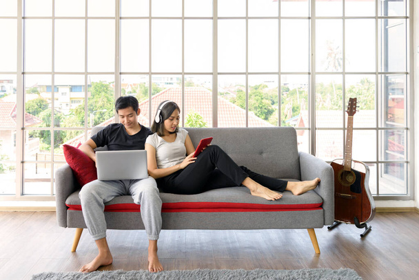 Nuori aasialainen pari käyttää rento vaatteita rentouttava istuu kodikas sohvalla olohuoneessa työskentelee tietokoneen kannettava tietokone ja tabletti PC kotona. Rakkaus, onnellisuus, työ kotona käsite - Valokuva, kuva