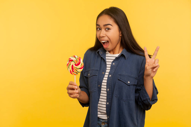 Sweet lollipop. Portrait of carefree joyful happy girl in denim shirt holding rainbow candy and showing victory or peace gesture, shouting excited. indoor studio shot isolated on yellow background - Foto, imagen