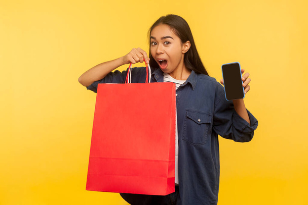 Portrait of thrilled amazed shopper girl in denim shirt holding bags and mobile phone with empty display mock up, surprised by crazy shopping in fashion store, thrift sale. indoor studio shot isolated - Zdjęcie, obraz