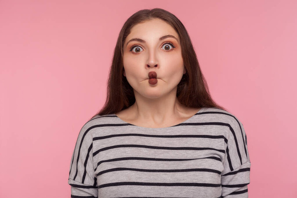 Portrait of funny wondered woman in striped sweatshirt making fish face grimace with lips and looking amazed confused with idiotic silly expression. indoor studio shot isolated on pink background - Foto, imagen