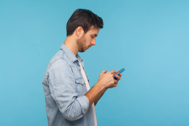 Side view of serious man in worker denim shirt texting message in social media on cell phone, using mobile network services, chatting online. indoor studio shot isolated on blue background, copy space - Fotografie, Obrázek