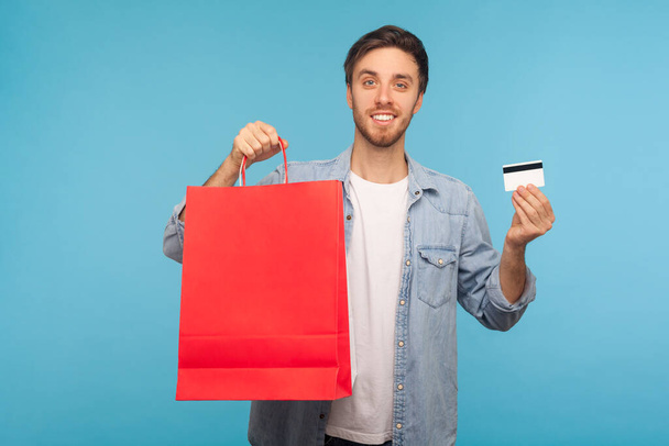 Portrait of joyful handsome man in stylish denim shirt holding credit card and shopping bags with blank space for advertise text, making purchases for electronic money. studio shot, blue background - Zdjęcie, obraz