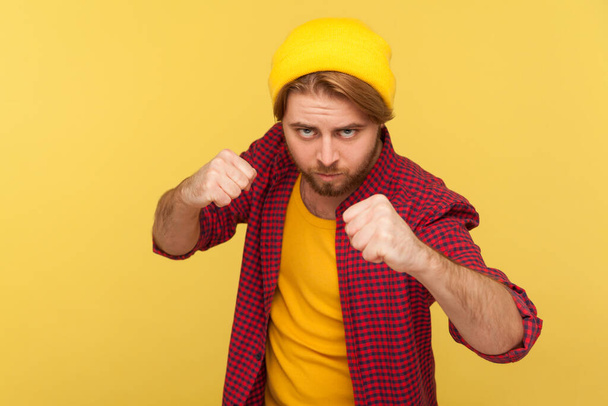 Aggressive hipster bearded guy in beanie hat, checkered shirt holding clenched fists up ready to boxing, punching with confident expression, fighting spirit. studio shot isolated on yellow background - Valokuva, kuva