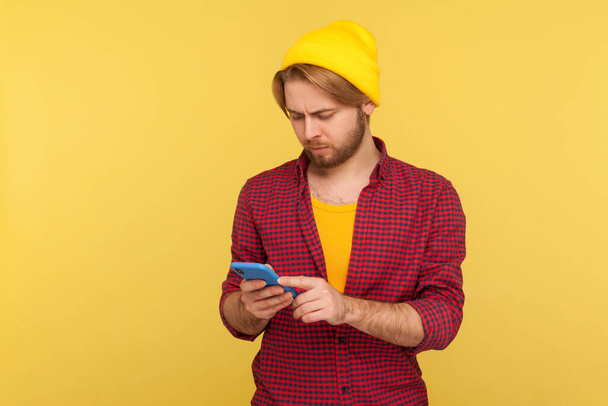 Hipster trendy guy in beanie hat and checkered shirt using cell phone, texting or reading message, chatting in social network, enjoying mobile application. studio shot isolated on yellow background - 写真・画像