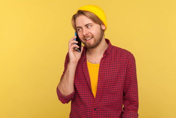 Cheerful trendy guy in hipster beanie hat and checkered shirt calling, smiling while talking mobile phone, having positive conversation on telephone. indoor studio shot isolated on yellow background - Foto, Bild