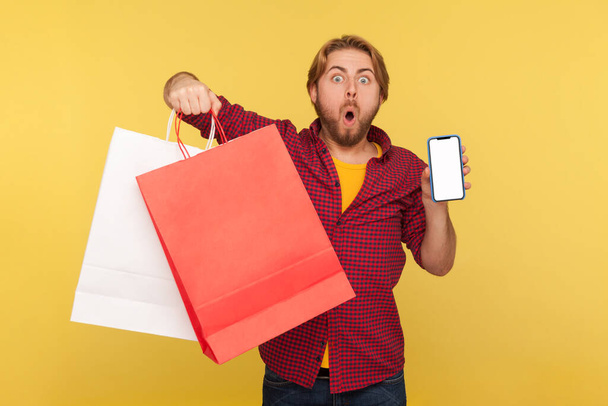 Amazed handsome shopper guy in checkered shirt holding shopping bags and mobile phone blank mock up display, looking at camera with shocked surprised expression. indoor studio shot isolated on yellow - Fotó, kép