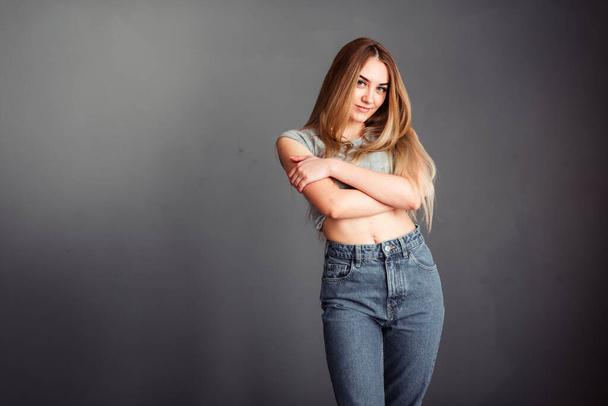 Photo portrait of a beautiful woman girl crossed her arms on her chest on a gray background in jeans with long blonde beautiful hair. Standing in front of a camera with a serious face - Fotoğraf, Görsel
