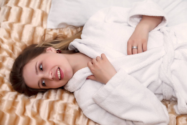 Girl of Slavic appearance in white coat on bed in hotel room. Pretty female shows acting emotions. Young charming woman is sitting on sofa in bedroom and playing with her feelings. Relax concept - Foto, Bild