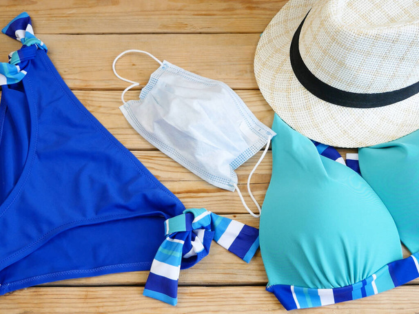 traje de baño de mujer, sombrero de sol y máscara médica sobre fondo de madera, Viajar en el mar durante el concepto pandémico coronavirus
 - Foto, Imagen