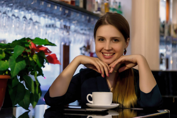 Girl of Slavic appearance in bar, cafe or restaurant with white Cup. Female shows emotion. Young charming woman is sitting at bar and drinking coffee or tea in her free evening time - Foto, afbeelding