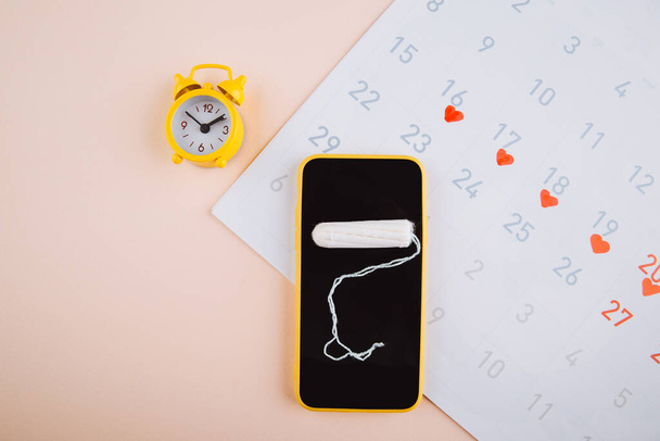 Smartphone sledovat svůj menstruační cyklus a pro značky. PMS a koncept kritických dnů. Bavlněný tampón a žlutý alarm na růžovém pozadí - Fotografie, Obrázek