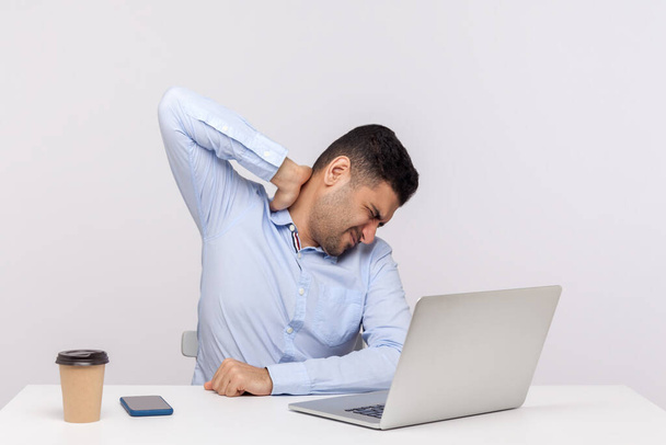 疲れた男性従業員はオフィスの職場に座って、背中の首の痛みに触れ、肩を傷つけるマッサージ、痛みを伴う硬い筋肉、疲労感。白い背景に隔離された屋内スタジオで - 写真・画像