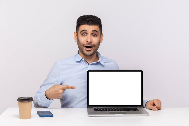 Wow, look at advertisement! Amazed man employee sitting in office workplace, pointing laptop blank display and looking surprised shocked expression, mock up for website promotion. studio shot isolated - Фото, изображение