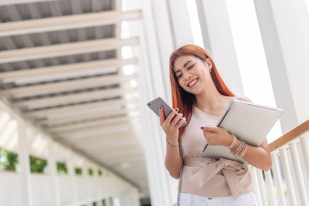 asiático joven hermosa mujer de negocios feliz sonriente celebración smartphone y portátil en moderno al aire libre oficina
. - Foto, Imagen