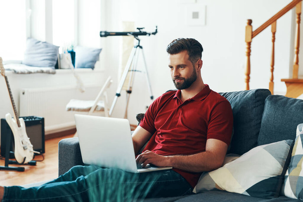 Handsome young man in casual clothing using laptop and smiling while sitting on the sofa - Foto, afbeelding