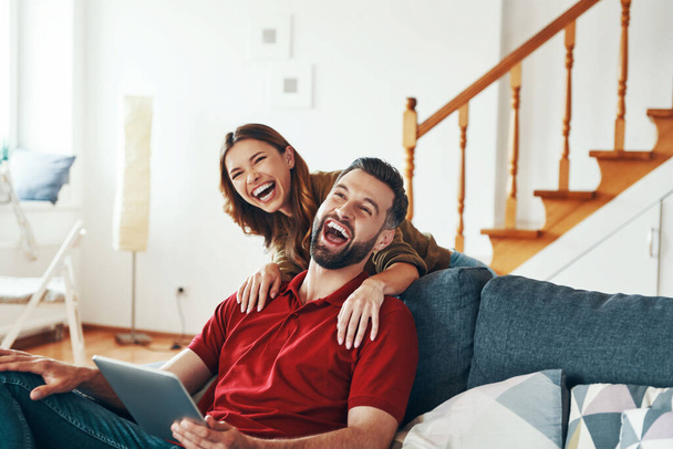 Cheerful young couple in casual clothing bonding together and laughing while resting on the sofa indoors    - Foto, imagen