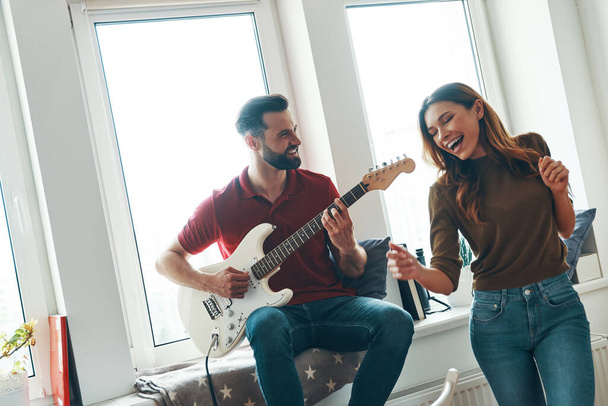 Happy young man in casual clothing playing the guitar while his attractive girlfriend dancing    - Фото, зображення