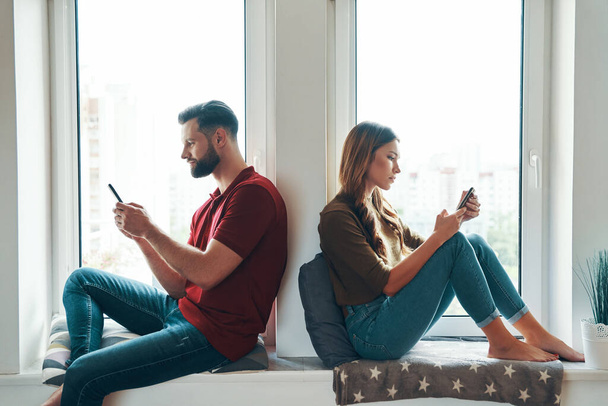 Busy young couple in casual clothing using smartphones while sitting on the window sill indoors             - Foto, Bild