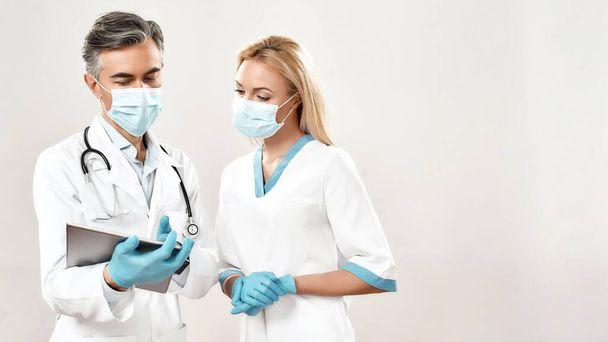 Δύο άνδρες και γυναίκες γιατροί με ιατρικές στολές και προστατευτικές μάσκες συζητούν το αποτέλεσμα ή τη διάγνωση ενώ στέκονται πάνω σε γκρι φόντο - Φωτογραφία, εικόνα