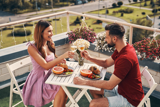 Top view of loving young couple in casual clothing toasting each other and smiling while sitting on the rooftop patio outdoors    - Fotografie, Obrázek