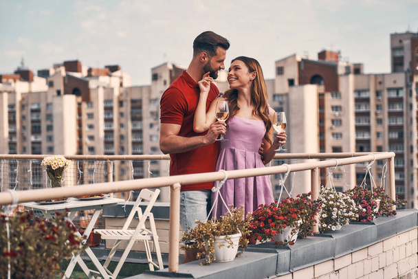 Charming young couple in casual clothing embracing and looking away with smile while standing on the rooftop patio outdoors    - Foto, immagini