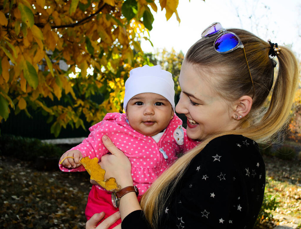 Młoda piękna szczęśliwa kobieta trzyma słodkie dziecko na zewnątrz w jesiennym parku. Młoda matka z trzymiesięczną córeczką w ramionach. Mieszana więź rodzinna. Blondynka śmiejąca się i urocze niemowlę patrzące w kamerę. - Zdjęcie, obraz