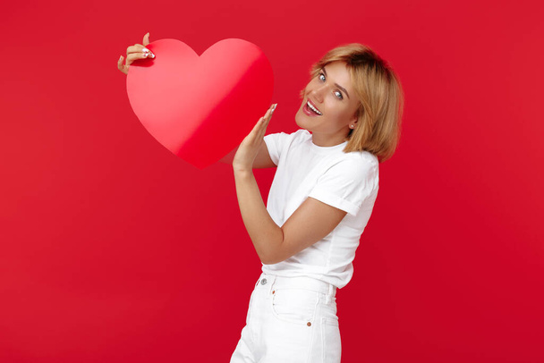 Positive young blonde woman in casual white outfit holding paper heart over red background. - Φωτογραφία, εικόνα