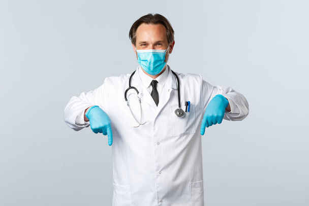 Covid-19, preventing virus, healthcare workers and vaccination concept. Pleasant doctor in medical mask and gloves give advice, recommend promo, pointing fingers down at banner - Photo, Image