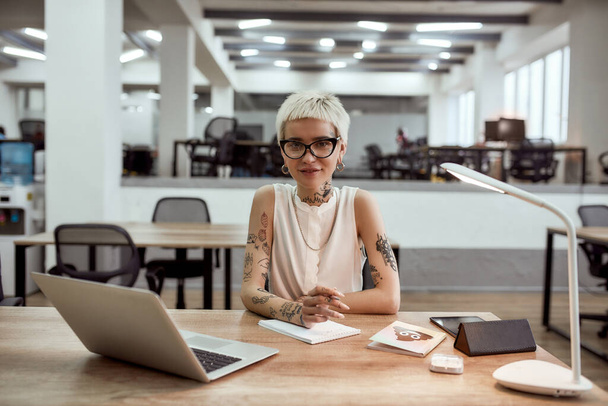 Stylish business woman. Young, positive tattooed business lady in eyewear looking at camera and smiling while working in the modern office - Zdjęcie, obraz