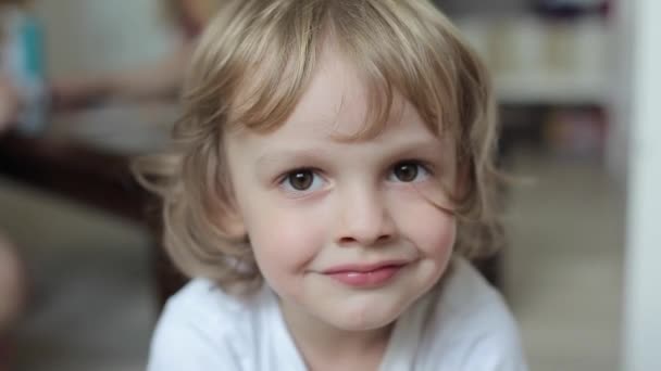 Retrato de un encantador niño rubio sonriente con el pelo rizado y con grandes ojos marrones en una camiseta blanca. Primer plano
 - Metraje, vídeo