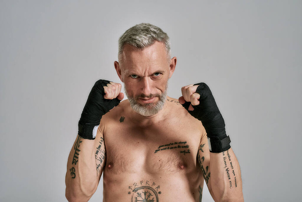 Half naked middle aged athletic man, kickboxer looking angry holding hands standing in studio over grey background. Muay Thai, Boxing or Kickboxing concept - Photo, image