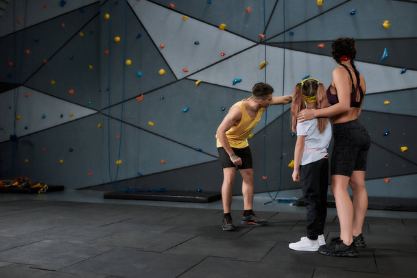 Full length shot of active little girl going to climb, male and female instructor helping kid, standing against artificial training climbing wall - Фото, зображення