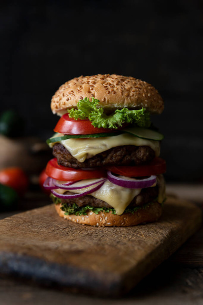 Hamburger with tomatoes, onions, cucumber, lettuce and melting cheese served on a  rustic wooden board  - Foto, Imagen