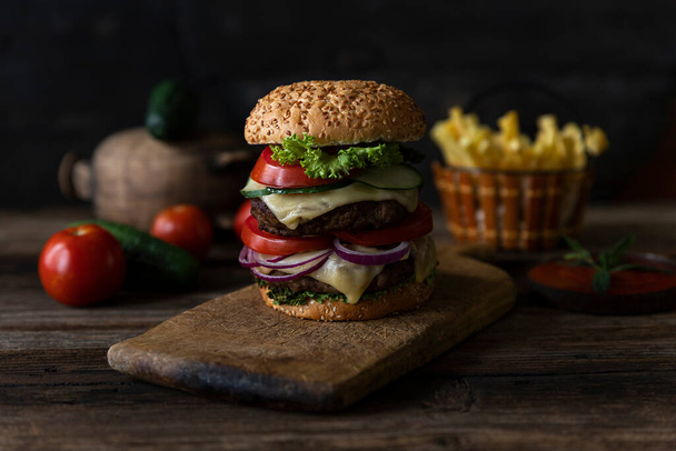 Hamburger with tomatoes, onions, cucumber, lettuce and melting cheese served on a  rustic wooden board with french fries and vegetables in background - Fotó, kép