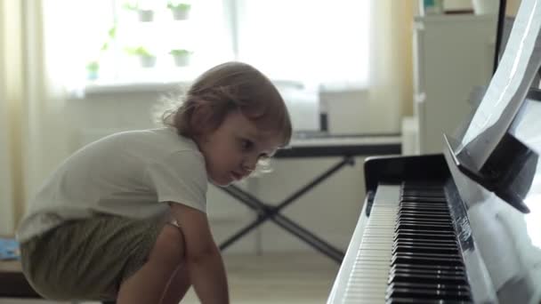Een blond jongetje met krullen en grote bruine ogen klimt op een stoel en drukt zachtjes op de pianotoetsen. Close-up - Video