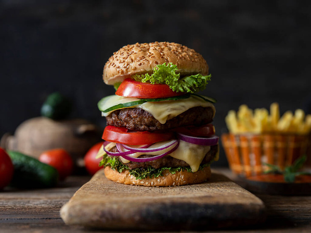 Hamburger with tomatoes, onions, cucumber, lettuce and melting cheese served on a  rustic wooden board with french fries and vegetables in background - Photo, image