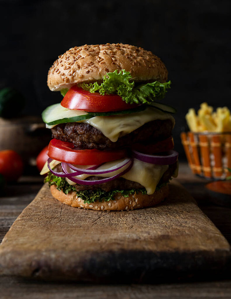 Hamburger with tomatoes, onions, cucumber, lettuce and melting cheese served on a  rustic wooden board with french fries and vegetables in background - Фото, изображение
