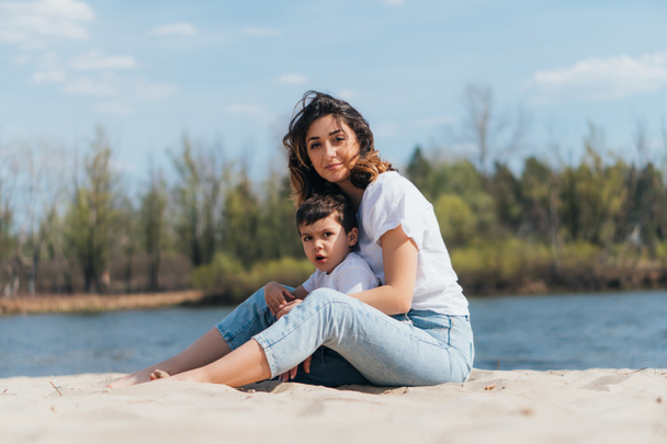 mother and son sitting on sand near river  - Foto, Imagem