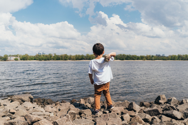 back view of cute boy holding rock near blue river  - Foto, Imagem