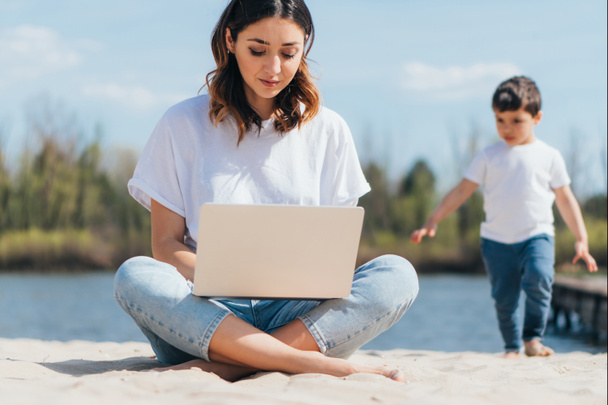 selective focus of freelancer using laptop while sitting on sand near cute son  - Fotoğraf, Görsel