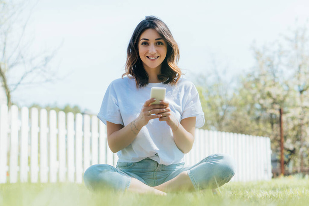 selective focus of cheerful woman sitting on green grass and holding smartphone - Valokuva, kuva