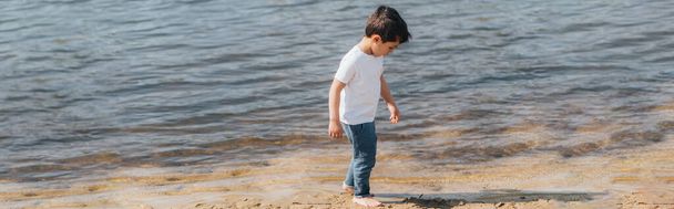 panoramic orientation of cute boy in denim jeans standing near lake  - Photo, image