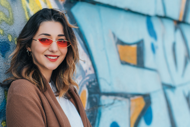 стильна жінка в сонцезахисних окулярах посміхається і стоїть біля стіни з графіті
 - Фото, зображення