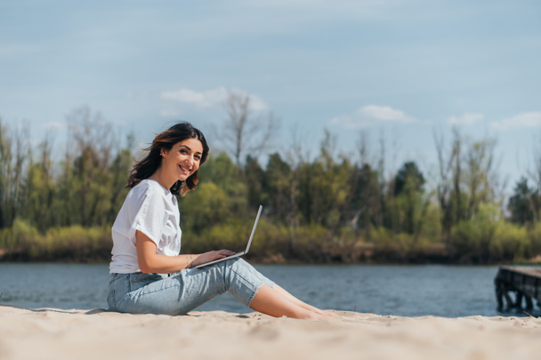 selective focus of happy freelancer using laptop near lake - Φωτογραφία, εικόνα