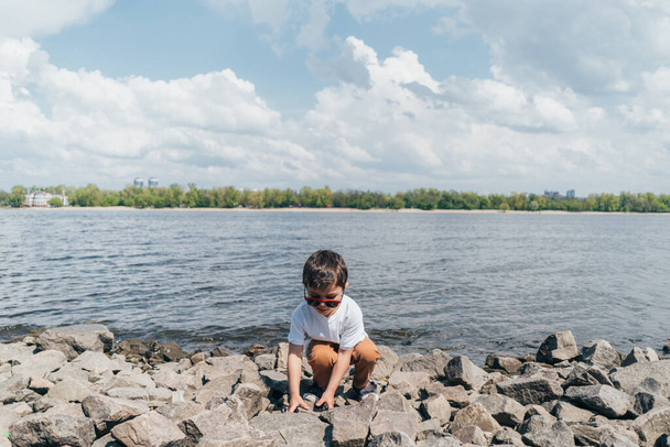 стильный мальчик в солнечных очках трогает камень, сидя у озера
 - Фото, изображение