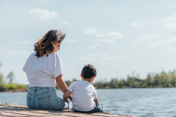 schöne Mutter schaut Sohn an, während sie am Fluss sitzt  - Foto, Bild