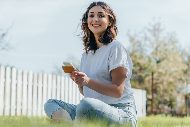 selective focus of cheerful woman sitting on grass, holding credit card and smartphone while online shopping  - Foto, afbeelding